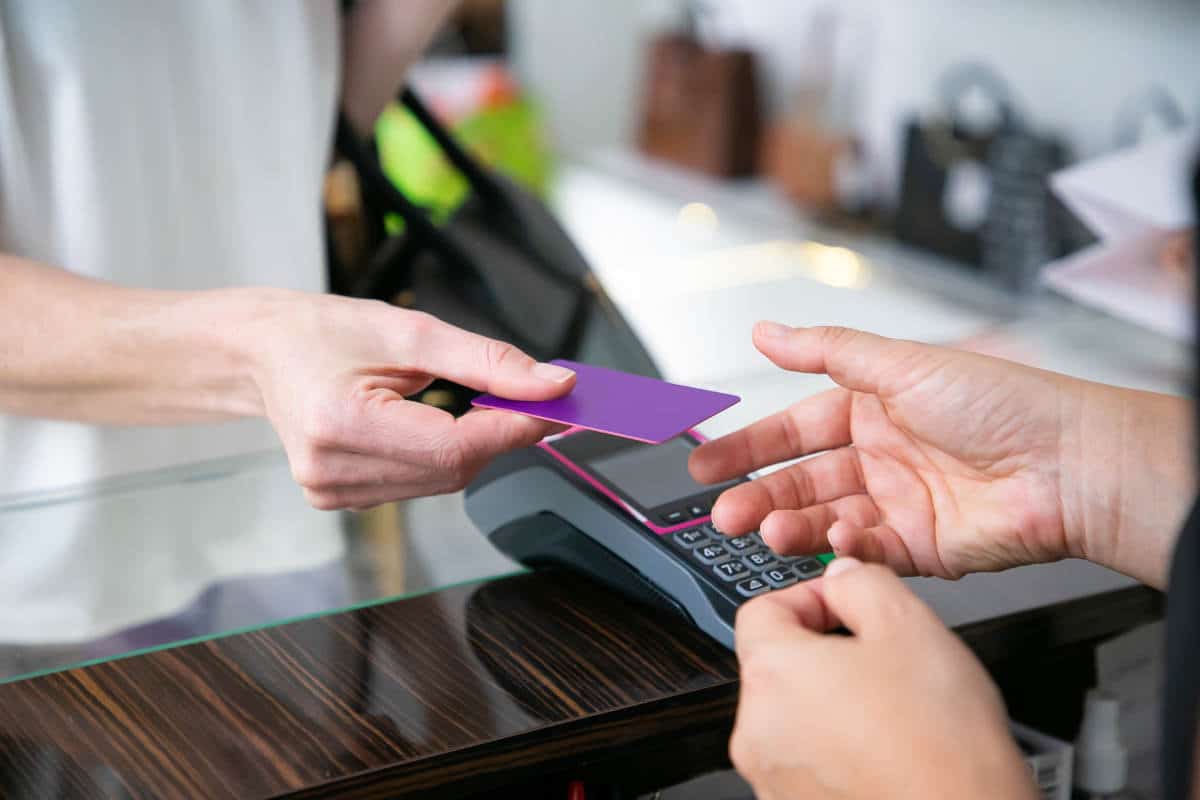 Mãos de um cliente entregando um cartão ao caixa sobre o balcão, ao lado de um terminal POS, em um contexto de compra.